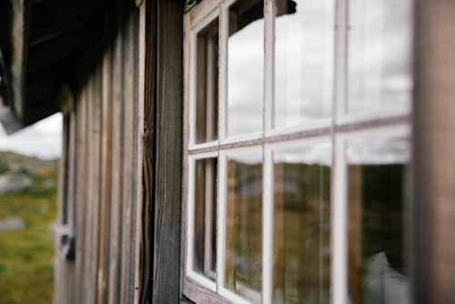 A close-up of a wooden building with a focus on a set of white-framed windows. The glass reflects an outdoor scene with soft, natural light, and the blurred background shows greenery and an overcast sky.