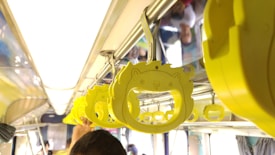 Bright yellow handles with a cartoonish animal design hang from a metal bar inside a public bus or train. Several passengers are visible in the background, with one person holding a handle.