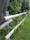 A wooden fence runs diagonally through a grassy area, bordered by dense green foliage and a large tree trunk. The fence is made of weathered logs and blends into the natural landscape.