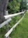 A wooden fence runs diagonally through a grassy area, bordered by dense green foliage and a large tree trunk. The fence is made of weathered logs and blends into the natural landscape.