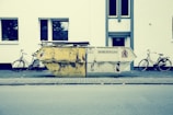 A large, worn-out dumpster is placed on a sidewalk in front of a white building. Two bicycles are parked on either side of the dumpster. The street scene appears calm and empty, with no people present. The building has simple, rectangular windows with reflections of trees.