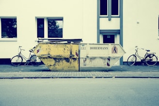 A large, worn-out dumpster is placed on a sidewalk in front of a white building. Two bicycles are parked on either side of the dumpster. The street scene appears calm and empty, with no people present. The building has simple, rectangular windows with reflections of trees.