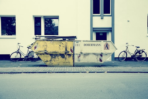 A large, worn-out dumpster is placed on a sidewalk in front of a white building. Two bicycles are parked on either side of the dumpster. The street scene appears calm and empty, with no people present. The building has simple, rectangular windows with reflections of trees.