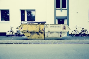 A large, worn-out dumpster is placed on a sidewalk in front of a white building. Two bicycles are parked on either side of the dumpster. The street scene appears calm and empty, with no people present. The building has simple, rectangular windows with reflections of trees.