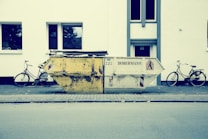 A large, worn-out dumpster is placed on a sidewalk in front of a white building. Two bicycles are parked on either side of the dumpster. The street scene appears calm and empty, with no people present. The building has simple, rectangular windows with reflections of trees.