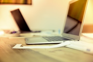 A modern workspace featuring two laptops placed on a wooden desk. The foreground shows a silver laptop with papers scattered nearby. In the background, another laptop is partially visible, slightly out of focus. The lighting gives a warm, inviting ambiance.