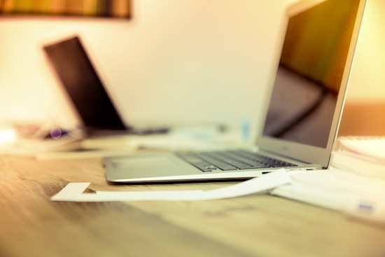 A modern workspace featuring two laptops placed on a wooden desk. The foreground shows a silver laptop with papers scattered nearby. In the background, another laptop is partially visible, slightly out of focus. The lighting gives a warm, inviting ambiance.