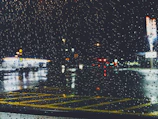 Rain-soaked street outside a glowing café window with blurred neon signs reflecting on wet pavement.