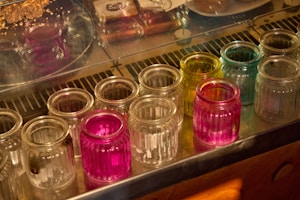 A collection of empty, ribbed glass jars arranged in rows on a reflective surface. Some jars are transparent, while others are colored in vibrant pink, yellow, and blue. The background features a display case with assorted baked goods.