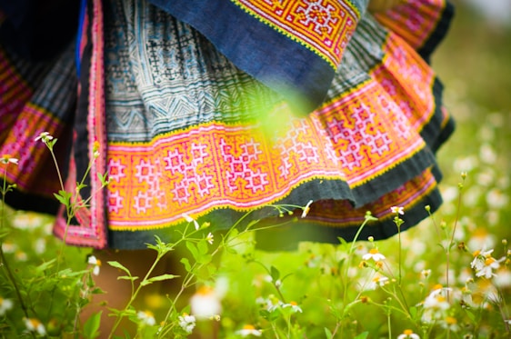 A colorful, intricately patterned skirt with vibrant pink, blue, and yellow embroidery is visible among a lush, green field of small white flowers. The fabric features geometric designs, and the scene conveys a sense of nature and traditional craftsmanship.