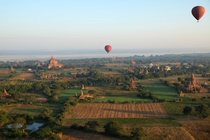 An expansive landscape featuring ancient structures spread across green fields and scattered trees. Several hot air balloons are floating above the terrain, and a river is visible near the horizon. The early morning sunlight casts a warm glow on the temples.