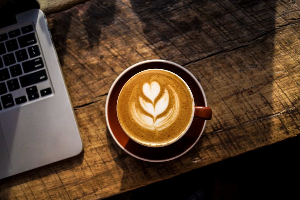caffe latte no copo de cerâmica branca ao lado de computador portátil prateado e preto