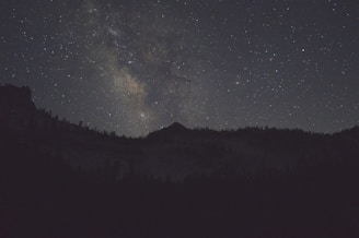 A star-filled night sky with a view of the Milky Way galaxy illuminated above the silhouette of a mountain range. The landscape is dark, contrasting with the bright and colorful galaxy overhead.