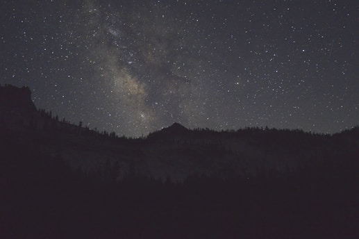 A star-filled night sky with a view of the Milky Way galaxy illuminated above the silhouette of a mountain range. The landscape is dark, contrasting with the bright and colorful galaxy overhead.
