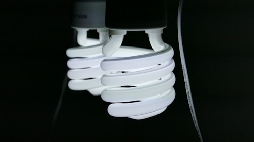 Two spiral-shaped fluorescent light bulbs emit a bright white glow against a dark background, with a visible power cord attached.