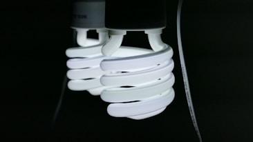 Two spiral-shaped fluorescent light bulbs emit a bright white glow against a dark background, with a visible power cord attached.
