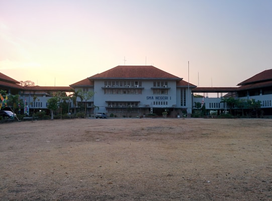 A school building with a large open courtyard in front, featuring a central structure with a red-tiled roof. It is flanked by two additional buildings with similar architecture on either side. The sky above is clear with a soft, warm glow, suggesting an early morning or late afternoon setting. Small trees and shrubs are situated near the buildings.