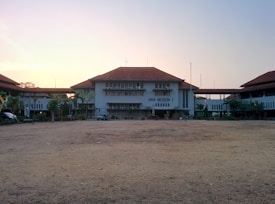 A school building with a large open courtyard in front, featuring a central structure with a red-tiled roof. It is flanked by two additional buildings with similar architecture on either side. The sky above is clear with a soft, warm glow, suggesting an early morning or late afternoon setting. Small trees and shrubs are situated near the buildings.