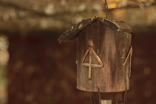A wooden birdhouse with a rustic appearance hangs against a blurred background. Its dark brown texture and natural wear give it an aged, earthy aesthetic. The birdhouse features geometric wooden decorations attached to its surface.