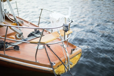 A close-up view of a wooden boat deck with polished wood and metal railing, floating on calm water. The ropes are neatly coiled, and there is a marine device attached to the railing. The water appears slightly rippled.