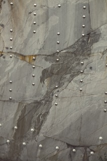 A pattern of smooth, metallic rivets arranged systematically on a textured, gray stone surface with visible natural lines and variations in color. The rivets reflect light, contrasting with the matte stone backdrop.