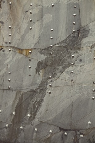 A pattern of smooth, metallic rivets arranged systematically on a textured, gray stone surface with visible natural lines and variations in color. The rivets reflect light, contrasting with the matte stone backdrop.
