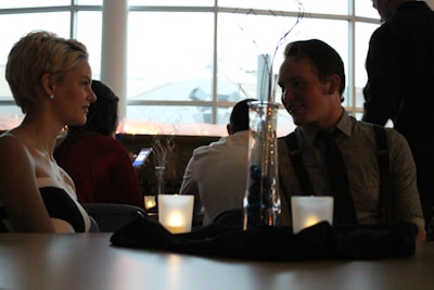 A dimly lit dining setting with a young woman and man sitting across from each other at a table. The woman has short blonde hair and is wearing earrings, while the man is dressed in a shirt and suspenders. Lit candles and a decorative glass vase sit on the table, and there are several people in the background, including someone using a phone. Large windows create a backdrop with soft, natural light.