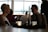 A dimly lit dining setting with a young woman and man sitting across from each other at a table. The woman has short blonde hair and is wearing earrings, while the man is dressed in a shirt and suspenders. Lit candles and a decorative glass vase sit on the table, and there are several people in the background, including someone using a phone. Large windows create a backdrop with soft, natural light.