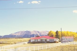 A rural scene features a charming farmhouse with a red roof surrounded by a white picket fence. In the background, majestic mountains stretch across the horizon under a clear blue sky. The landscape is adorned with vibrant autumn trees displaying yellow foliage.