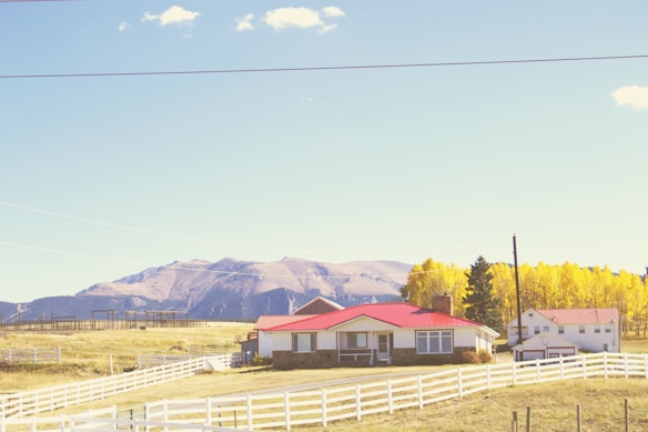 A rural scene features a charming farmhouse with a red roof surrounded by a white picket fence. In the background, majestic mountains stretch across the horizon under a clear blue sky. The landscape is adorned with vibrant autumn trees displaying yellow foliage.