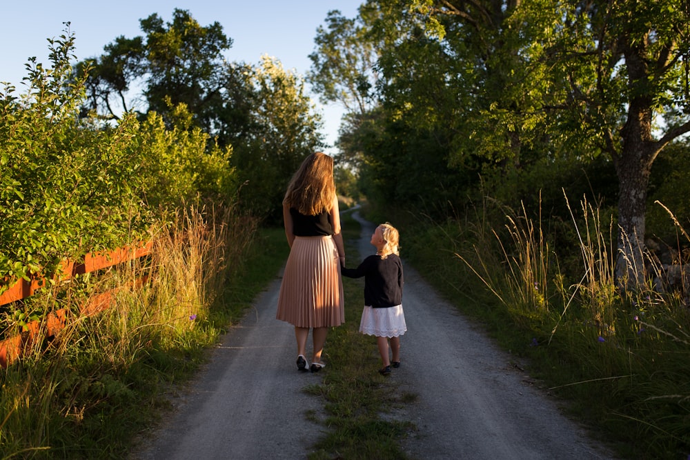 woman and girl walking on road surrounded by green grass