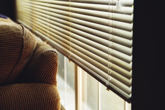 A patterned fabric armrest of a sofa is positioned beside a window with white horizontal blinds. The blinds are partially open, allowing soft natural light to filter through, creating a warm and cozy atmosphere.