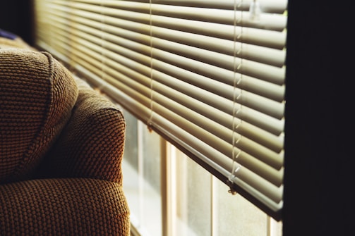A patterned fabric armrest of a sofa is positioned beside a window with white horizontal blinds. The blinds are partially open, allowing soft natural light to filter through, creating a warm and cozy atmosphere.