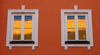 Two white-framed windows are set in an orange wall. The glass reflects a warm yellow hue with a hint of blue sky, possibly showing part of another building or structure. The window frames are detailed with decorative white moldings.