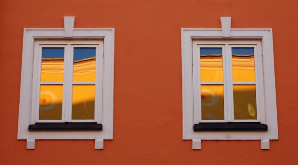 Two white-framed windows are set in an orange wall. The glass reflects a warm yellow hue with a hint of blue sky, possibly showing part of another building or structure. The window frames are detailed with decorative white moldings.