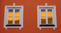 Two white-framed windows are set in an orange wall. The glass reflects a warm yellow hue with a hint of blue sky, possibly showing part of another building or structure. The window frames are detailed with decorative white moldings.