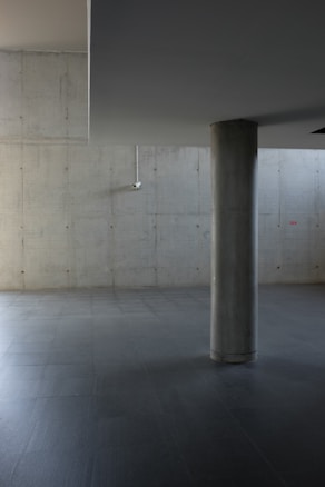 The image presents an interior space with a minimalist and industrial aesthetic. It features smooth concrete walls and flooring, with a large cylindrical column positioned slightly off-center. A small security camera is mounted on the upper left corner of the wall. The lighting is soft, casting subtle shadows, enhancing the muted color palette of the space.