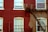 A brick building facade with three windows, two of which have white curtains. A fire escape staircase runs diagonally across the wall. The building has bricks painted in red, with some patches of grey where painting is incomplete.
