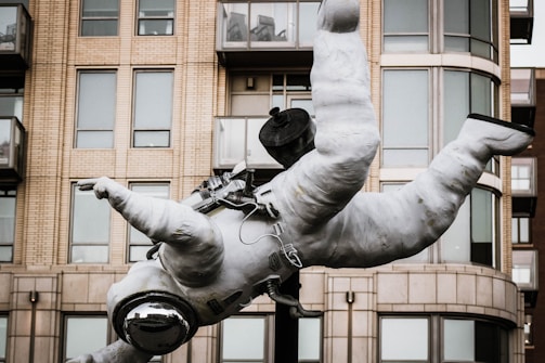 A sculpture of an astronaut is suspended mid-air in front of a modern residential building with brick and glass facades. The astronaut is wearing a full spacesuit, including a helmet with a reflective visor. The building in the background features multiple windows and several small balconies.