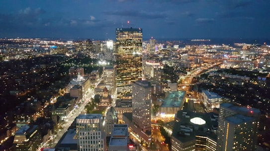 A cityscape during the evening with illuminated skyscrapers, bustling streets, and distant streetlights. The horizon reveals a darkening sky with clouds. The urban landscape is filled with a mix of modern and historic architecture, showing a vibrant and lively city atmosphere.