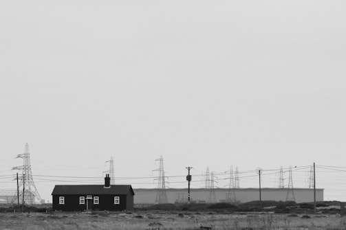 A small, dark-colored house with a chimney sits in a barren landscape. Several large electrical pylons stand in the background, connected by wires, with a flat, industrial-looking building behind them. The sky is overcast, creating a stark, monochrome appearance.