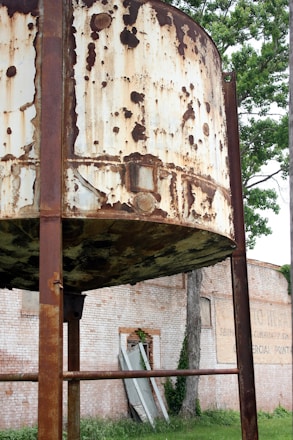 A large, rusty cylindrical metal structure supported by beams is prominent in the foreground. The metal surface is heavily corroded, displaying patches of rust and peeling paint. In the background, there is a brick wall with some foliage climbing up. Some planks and debris are scattered near the base of a tree visible against the wall.