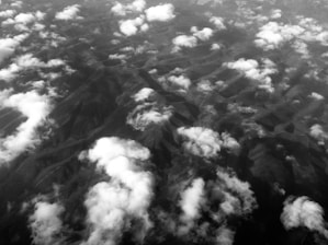 An aerial view displaying a landscape covered with scattered clouds. The terrain below appears to be mountainous, with a series of ridges and valleys. The image is in black and white, highlighting contrasts between clouds and land.