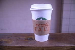 A white disposable coffee cup with a brown cardboard sleeve reading 'warmly, FALL', resting on a wooden surface. The cup has a green mermaid logo partially visible on the upper section. The background is blurred, highlighting the focus on the cup.