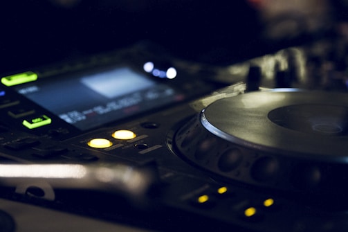 A close-up view of a DJ mixer with illuminated buttons and displays, featuring a large jog wheel and several control knobs. The lighting is dim and moody, highlighting the electronic equipment in a dark environment.