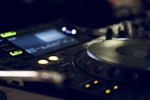A close-up view of a DJ mixer with illuminated buttons and displays, featuring a large jog wheel and several control knobs. The lighting is dim and moody, highlighting the electronic equipment in a dark environment.