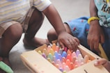 Two children are sitting on the floor, engaged in selecting colorful chalk pieces from a box. One child is wearing shorts and has a striped shirt, while the other wears a skirt with small embroidered cherries. The hands of both children appear to be reaching for the chalk, showing interaction and involvement in a creative activity.