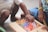 Two children are sitting on the floor, engaged in selecting colorful chalk pieces from a box. One child is wearing shorts and has a striped shirt, while the other wears a skirt with small embroidered cherries. The hands of both children appear to be reaching for the chalk, showing interaction and involvement in a creative activity.