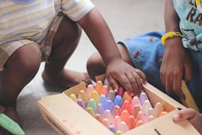 Two children are sitting on the floor, engaged in selecting colorful chalk pieces from a box. One child is wearing shorts and has a striped shirt, while the other wears a skirt with small embroidered cherries. The hands of both children appear to be reaching for the chalk, showing interaction and involvement in a creative activity.
