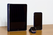 A tablet, smartphone, and smartwatch are arranged in a neat row on a wooden surface. The devices have black screens and metallic outlines, with a blue stand or case complementing each item. The background is a plain, light-colored wall.